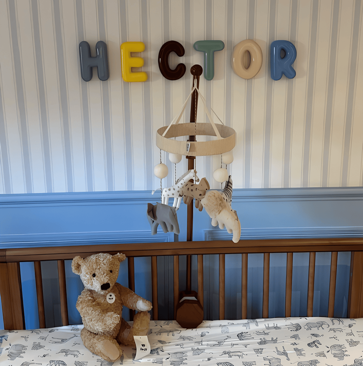 Nursery with a wooden crib, teddy bear, and mobile against a striped wall with 'HECTOR' ceramic letters.