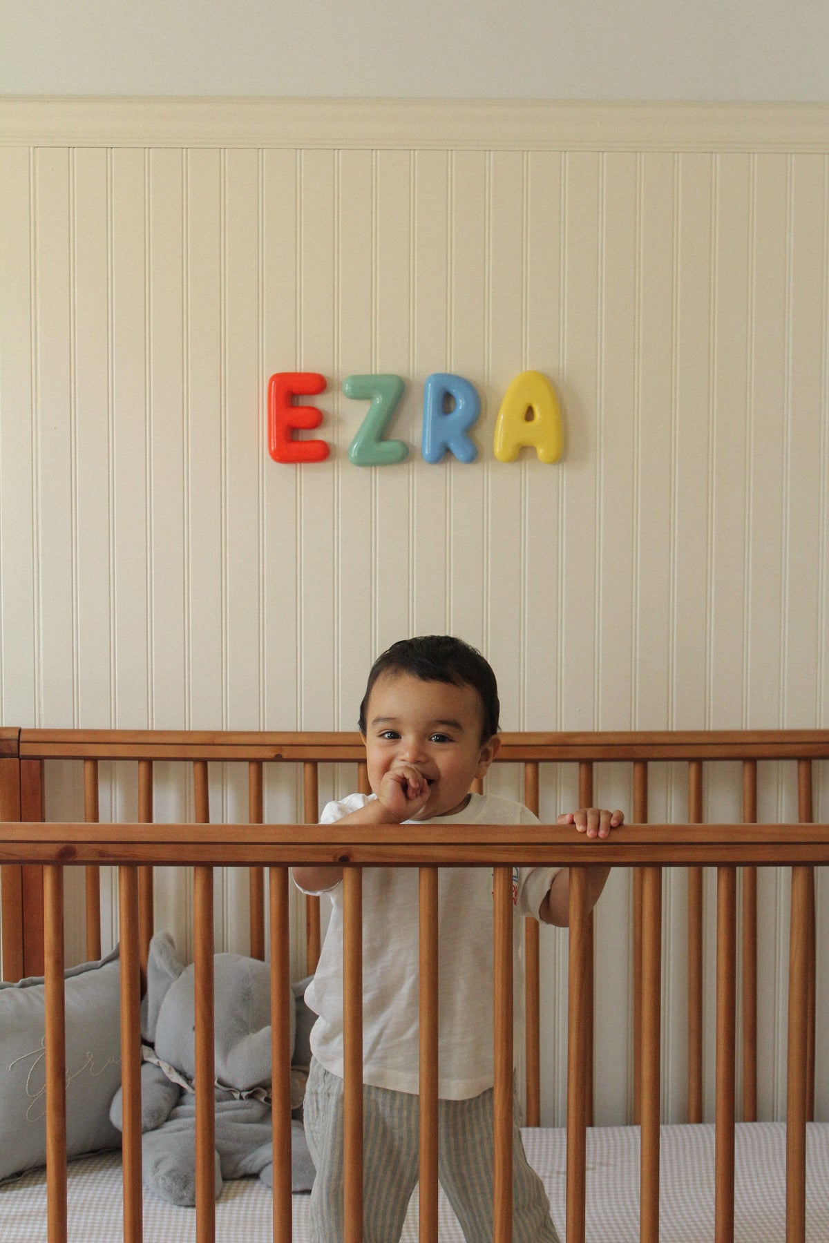 Child standing in a crib with colorful bubble letters spelling 'EZRA' on the wall.