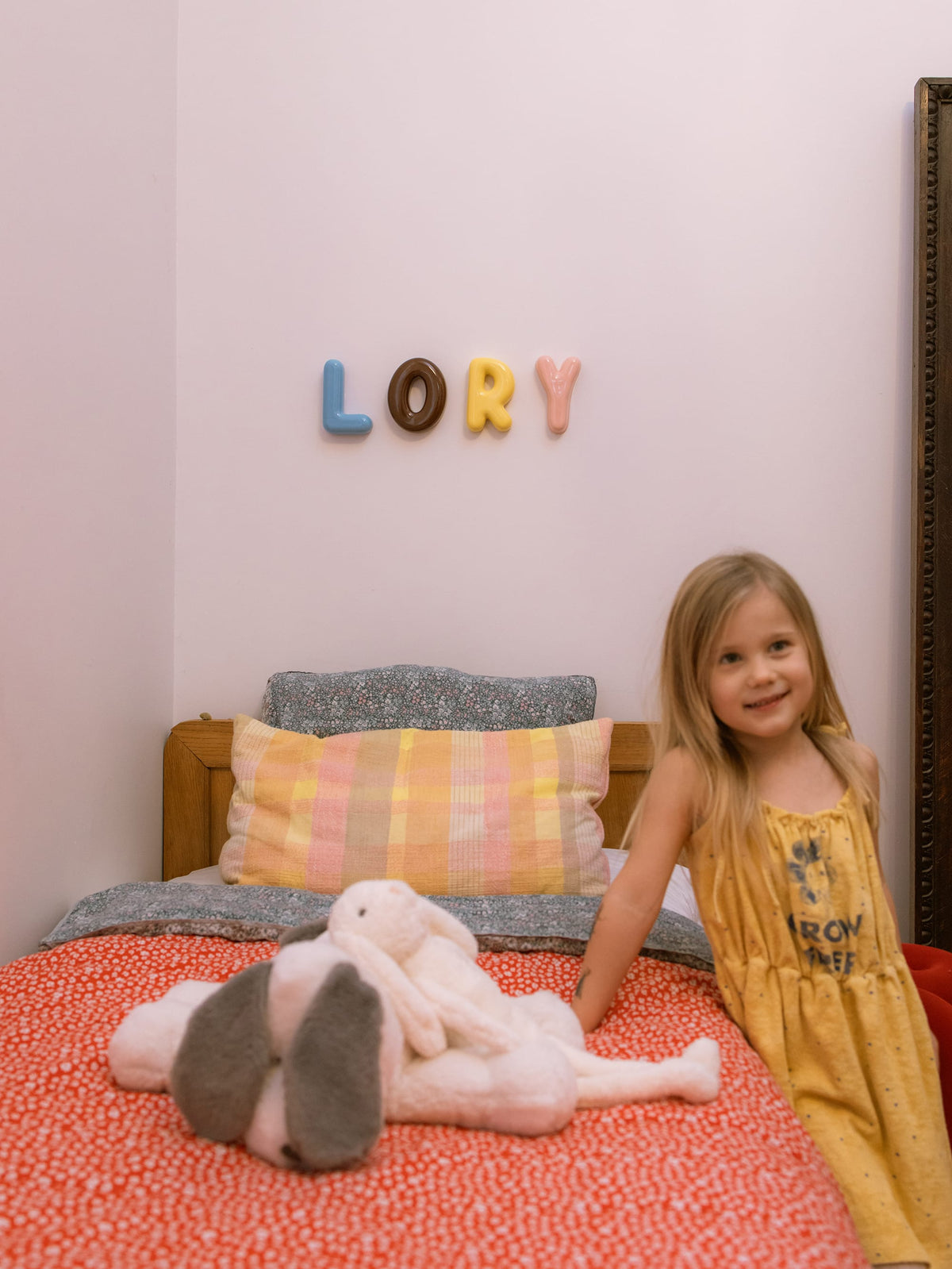 Young girl sitting on a bed with stuffed animals, colorful pillows, and 'LORY' ceramic letters on the wall.